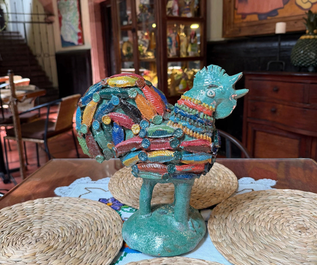 Primer plano de una pieza artesanal de un gallo de barro vidriado color verde de San José de Gracia, Michoacán, exhibido sobre una base de madera, con las vitrinas de la Artisans Collection y el pasillo del hotel Mansión Iturbe difuminados al fondo.
