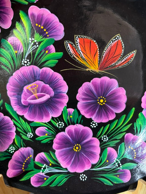 Acercamiento al detalle pintado a mano de una mariposa monarca y flores moradas sobre el fondo de madera negra de una batea artesanal de Santa Fe de la Laguna, Michoacán.