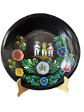 Vista frontal completa de una batea circular de madera negra pintada a mano con la Danza de los Viejitos al centro y flores tradicionales en tonos azul, blanco, amarillo y rosa alrededor.