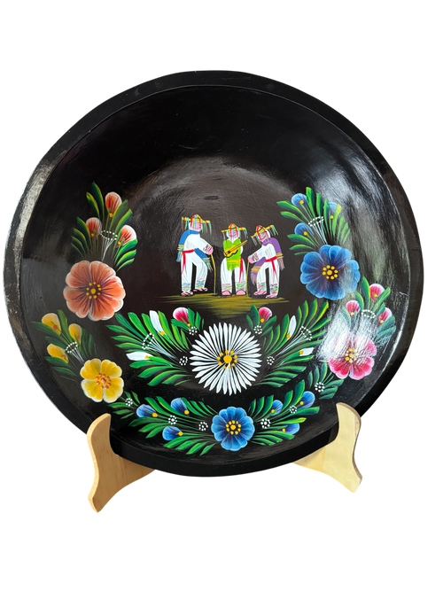 Vista frontal completa de una batea circular de madera negra pintada a mano con la Danza de los Viejitos al centro y flores tradicionales en tonos azul, blanco, amarillo y rosa alrededor.